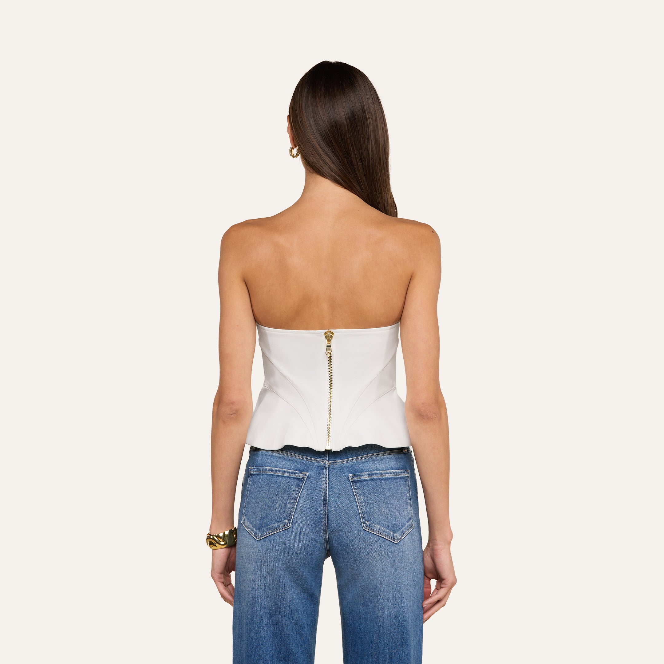 Una donna dai lunghi capelli castani lisci indossa il top a corsetto Anna — bianco, senza spalline, con una cerniera dorata e orlo smerlato — abbinato a jeans blu e gioielli dorati. Lo sfondo è beige e conferisce all’insieme un’atmosfera da alta moda.