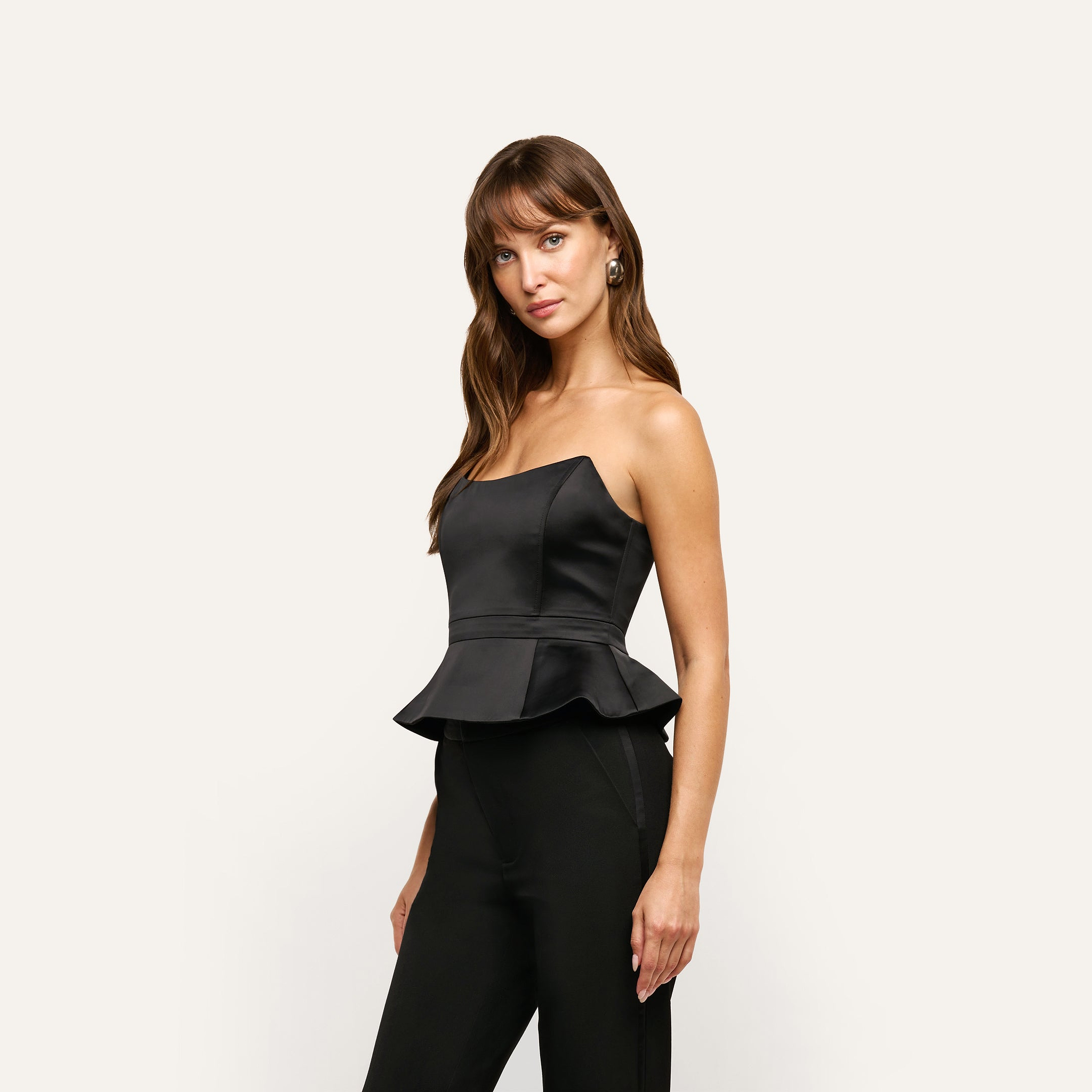A person wears a black strapless satin Meliora Corset Peplum Top and black trousers.