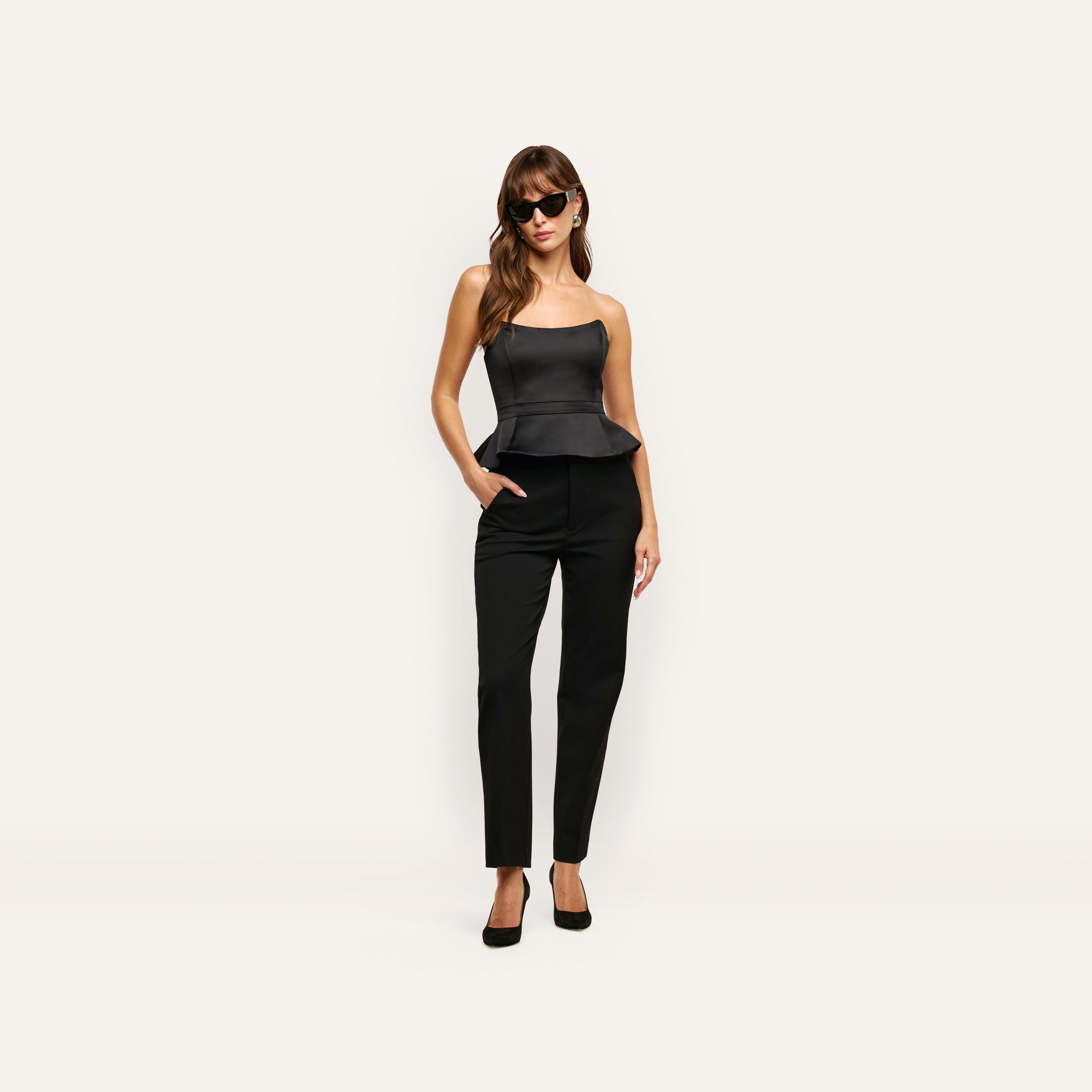 A woman wearing a black satin Meliora Corset Peplum Top with black trousers and heels.