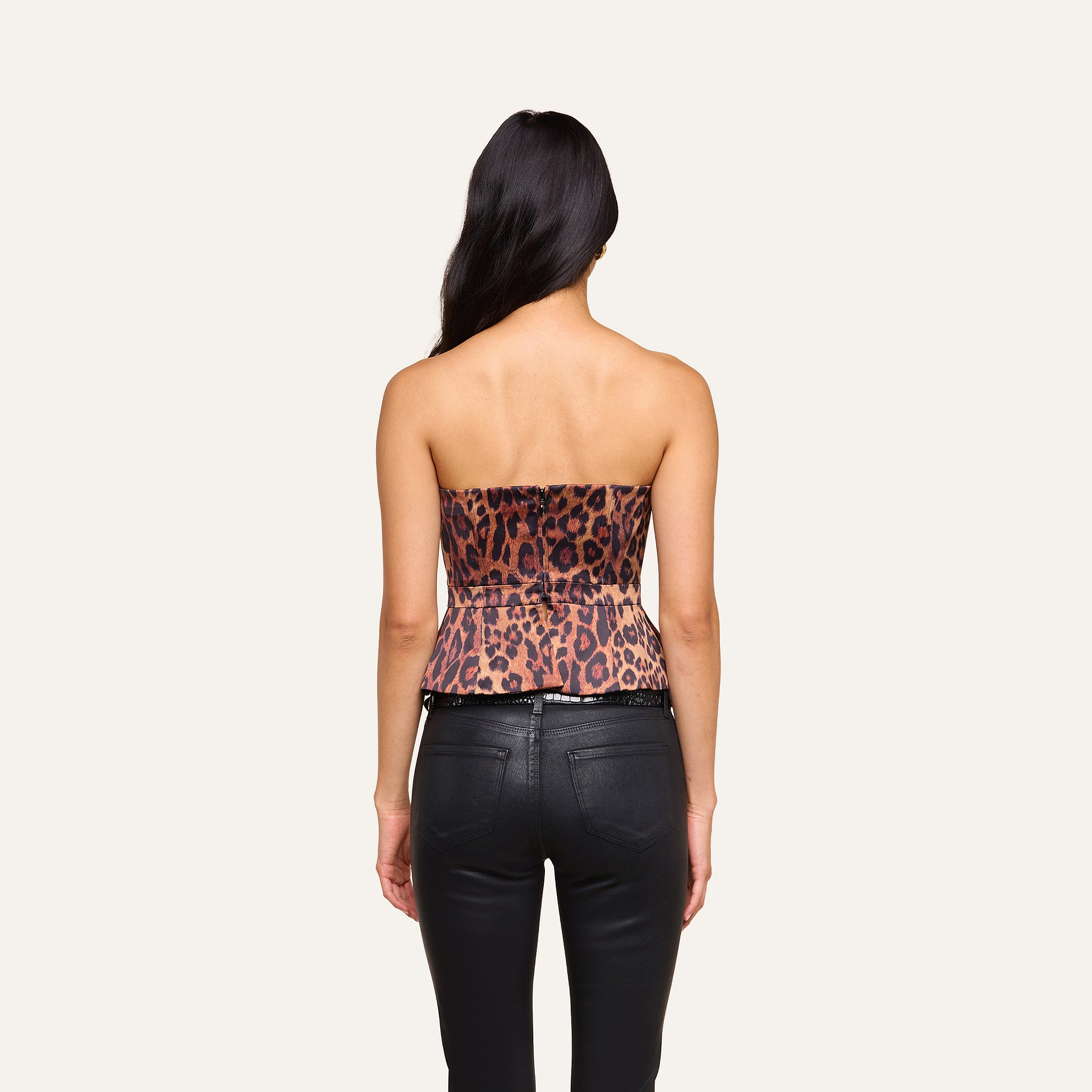 A woman with long dark hair stands with her back to the camera, wearing the Meliora Corset Peplum Top in a leopard print style and black leather pants against a plain light background.