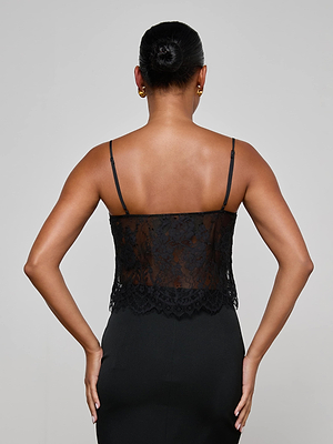 A woman faces away from the camera, wearing the L'AGENCE Caylee Lace Camisole with thin straps and a fitted black skirt. Her hair is styled in a neat bun, complemented by gold hoop earrings.