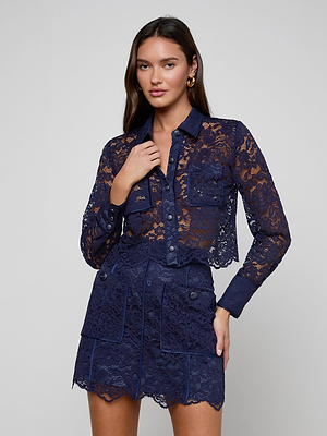 A woman with long brown hair and gold hoop earrings models the Roelle Lace Blouse—a sheer navy, long-sleeve button-up shirt with front pockets—paired with a matching high-waisted scalloped skirt against a plain light gray background.
