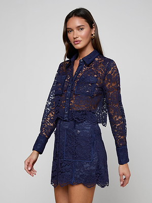A woman poses against a plain background wearing the Roelle Lace Blouse—a sheer navy, long-sleeve button-up—paired with a matching lace skirt featuring patch pockets and scalloped edges.