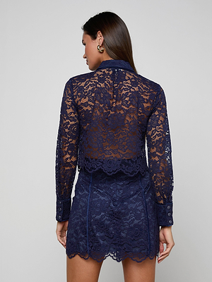 A woman faces away, wearing the Roelle Lace Blouse in sheer navy with long sleeves, gold buttons, and a scalloped hem, paired with a matching navy lace mini skirt that reveals glimpses of skin through the delicate fabric.
