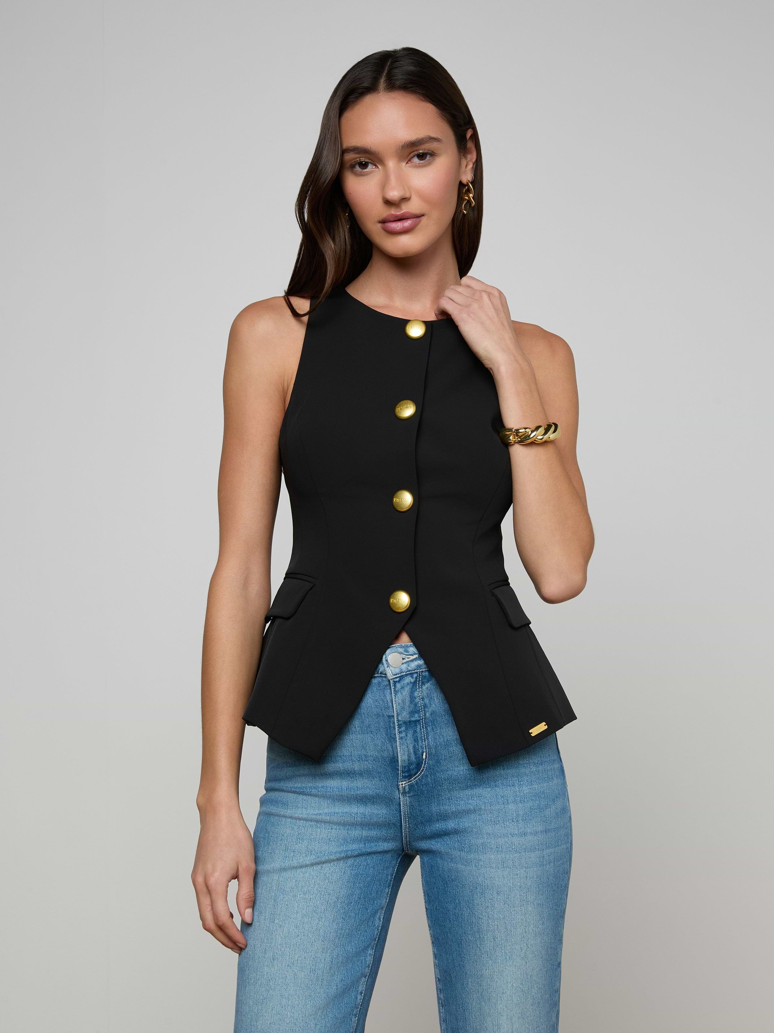 A woman wearing the Tessa Vest, a sleeveless black top with gold buttons, paired with blue jeans stands against a plain light background, touching her hair.