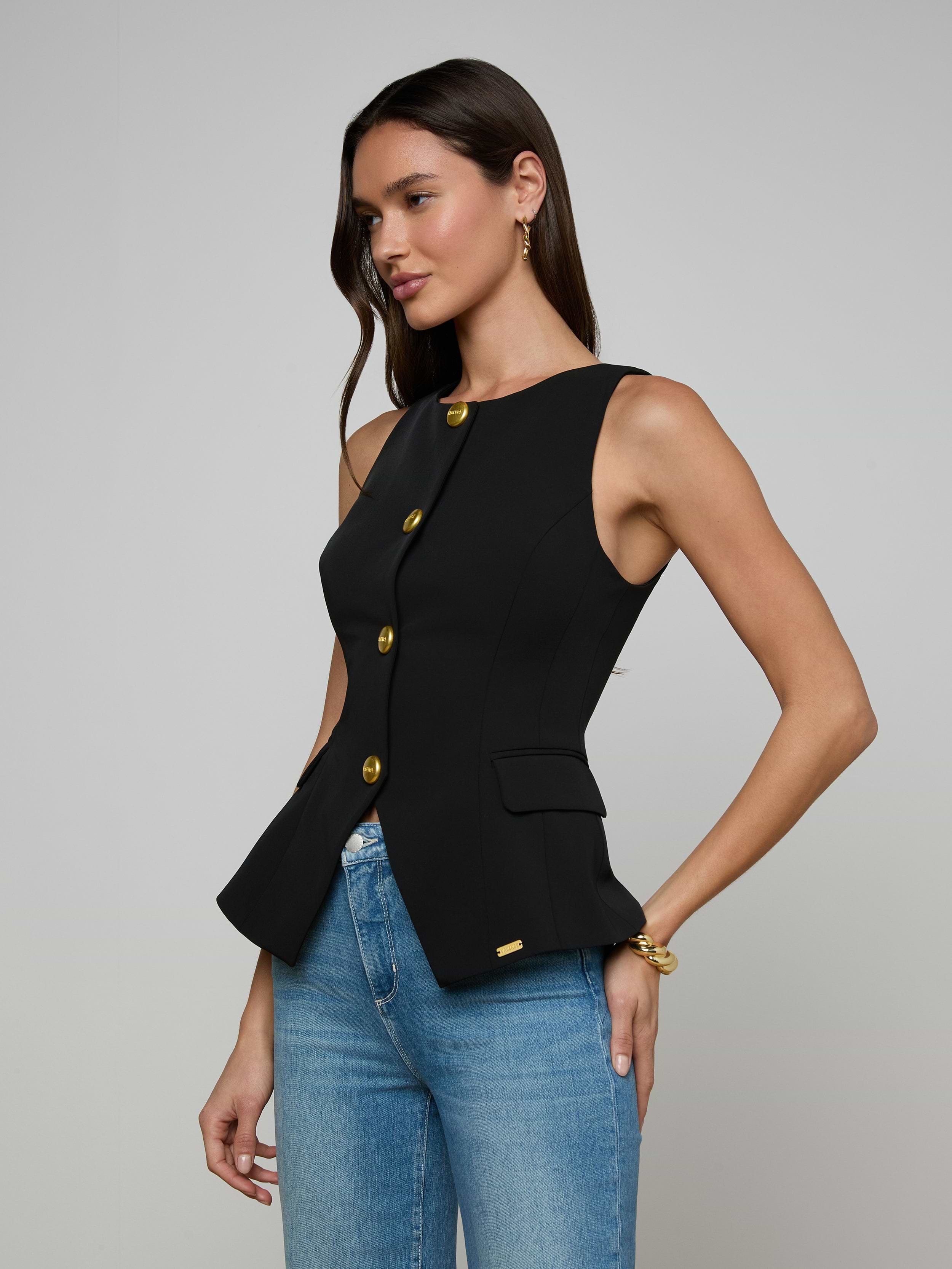 A woman wearing the Tessa Vest—a sleeveless suit vest with gold buttons—paired with blue jeans stands against a plain light background.