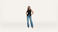 A woman stands against a white background wearing a sleeveless black top, blue jeans, and heeled sandals, with her hands in her pockets and a neutral expression.
