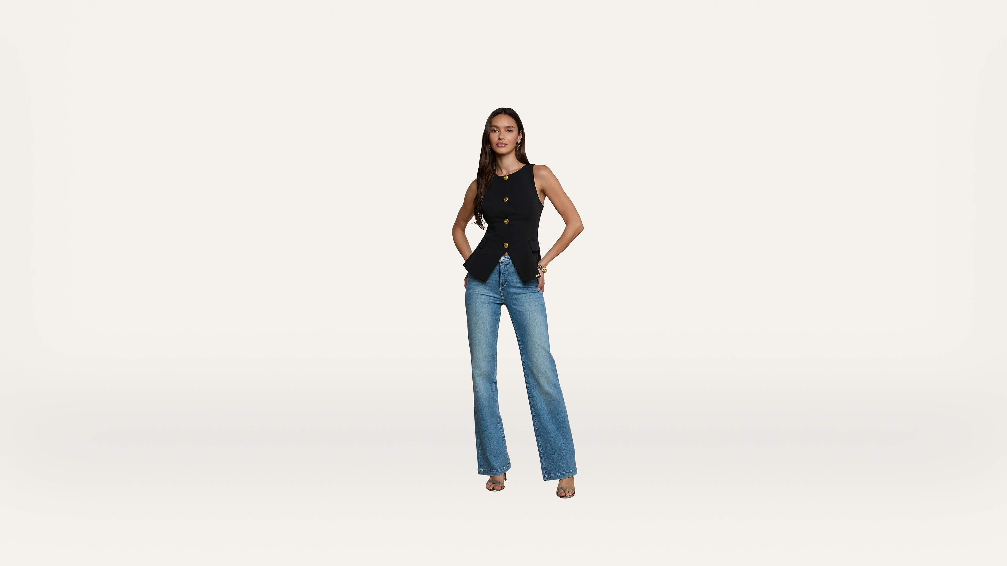 A woman stands against a white background wearing a sleeveless black top, blue jeans, and heeled sandals, with her hands in her pockets and a neutral expression.