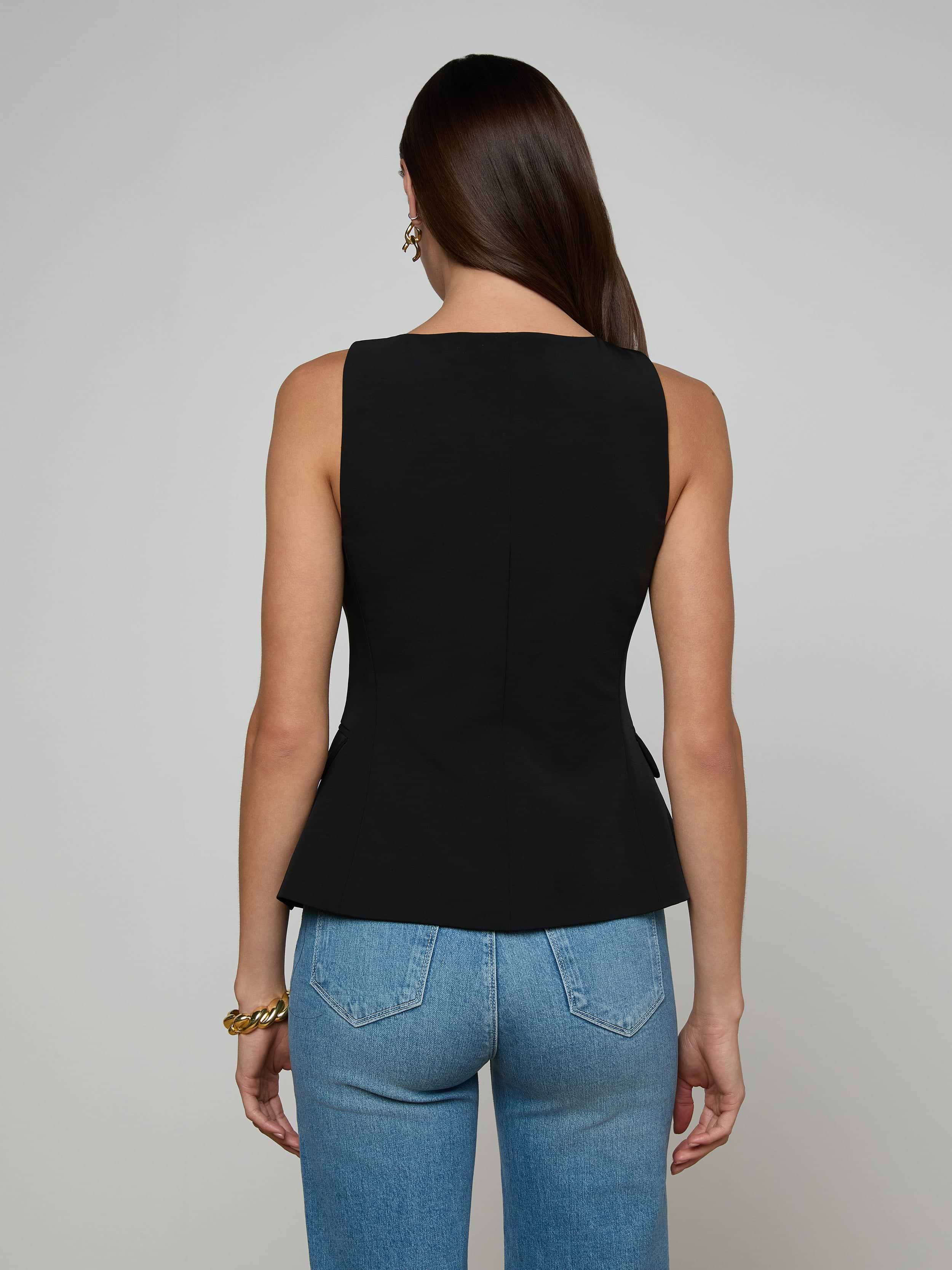 A woman with long brown hair is shown from the back, wearing the Tessa Vest—a sleeveless suit top with gold buttons—paired with blue jeans against a plain light background.