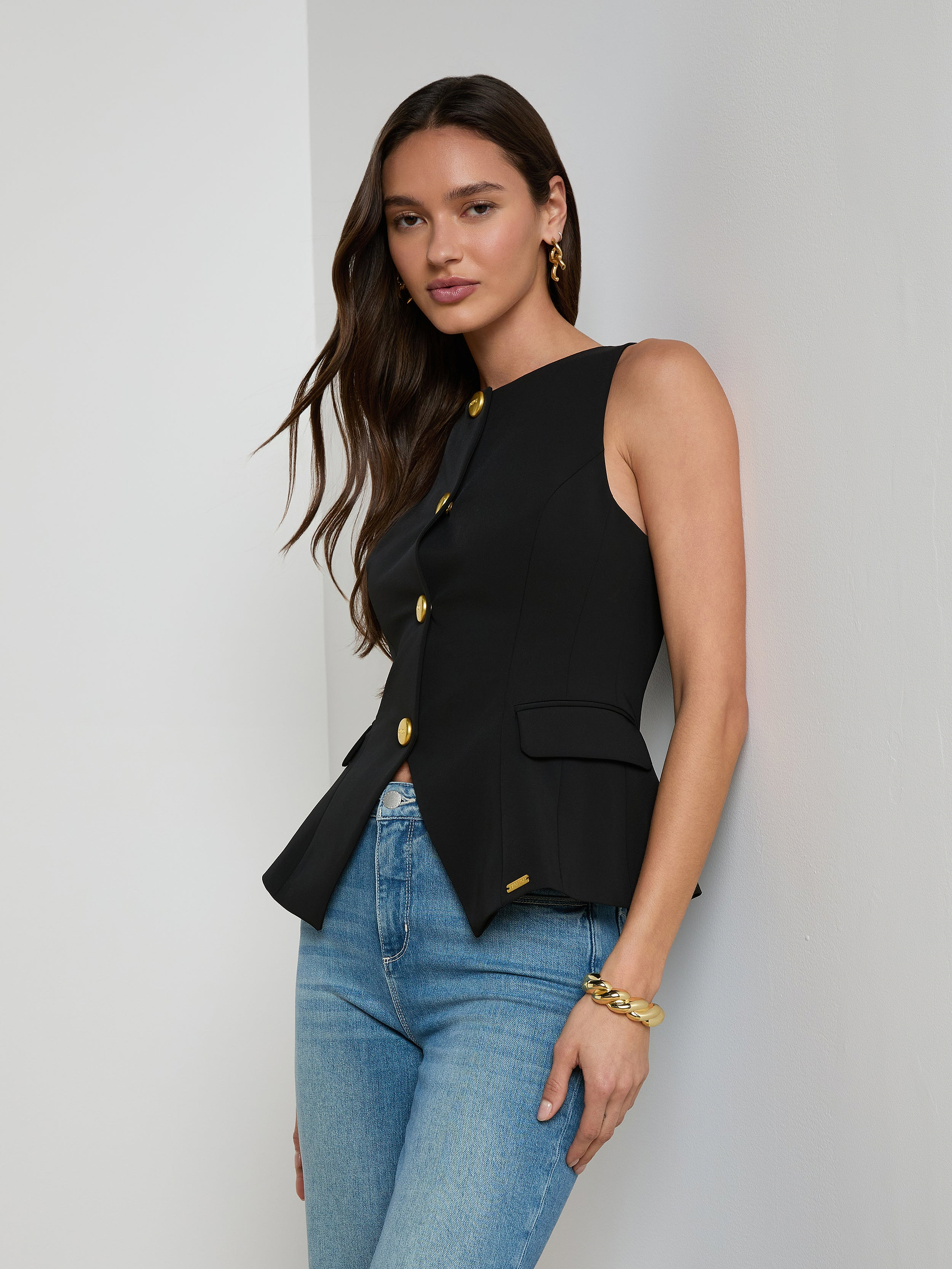 A woman with long brown hair models the Tessa Vest—a sleeveless, suit-style black vest crafted from stretch suit fabric and featuring gold buttons—paired with blue jeans and gold jewelry, standing against a plain light background.
