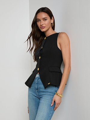 A woman with long brown hair wears the Tessa Vest, a sleeveless stretch suit top with gold buttons, paired with blue jeans and gold jewelry, standing against a white wall and looking at the camera.