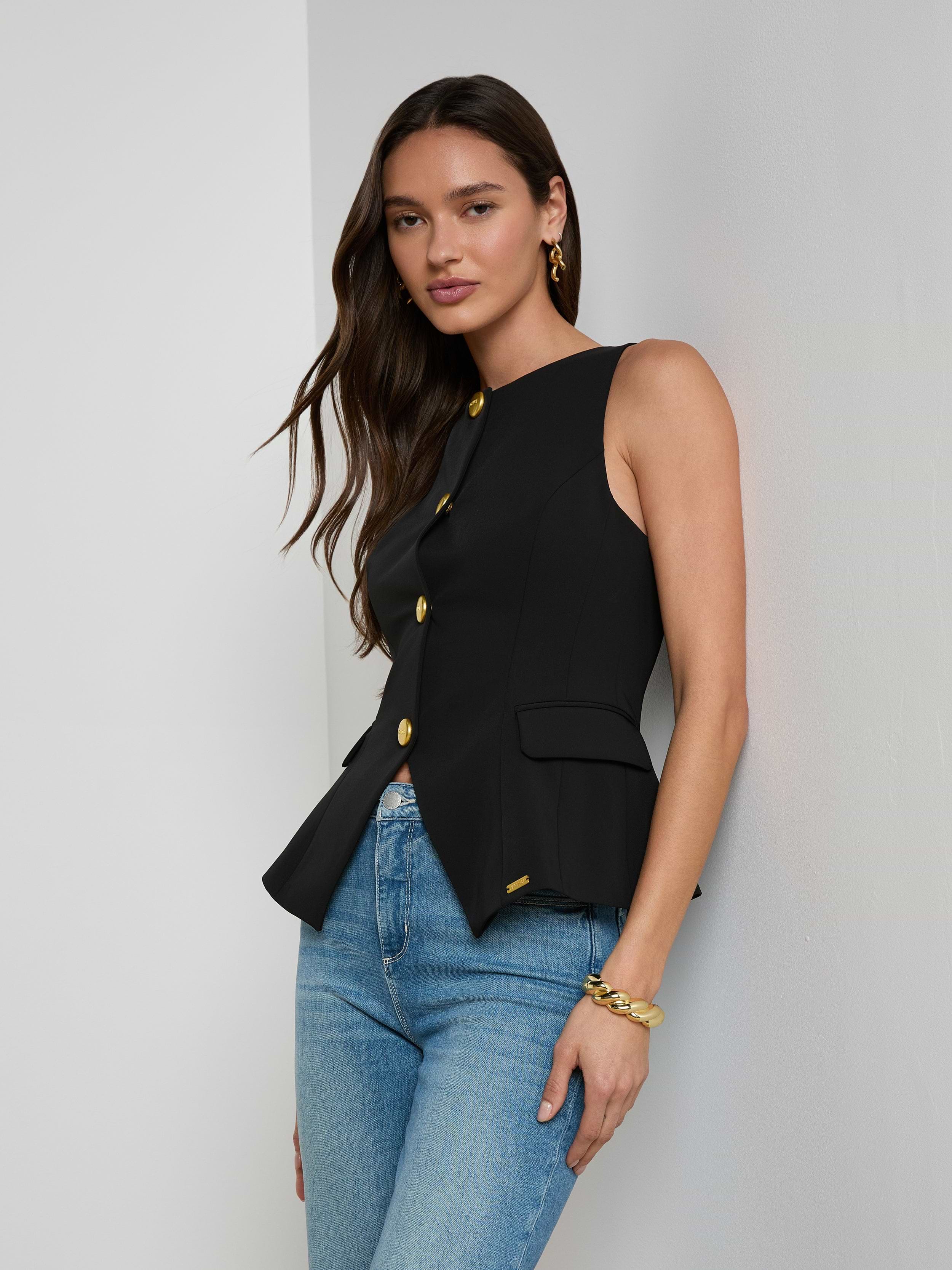 A woman with long brown hair models the Tessa Vest—a sleeveless, suit-style black vest crafted from stretch suit fabric and featuring gold buttons—paired with blue jeans and gold jewelry, standing against a plain light background.