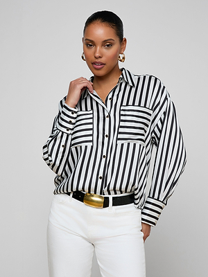 A woman poses confidently in the Eliora Shirt—a relaxed-fit striped button-down—paired with white pants, a black belt with a gold buckle, gold hoop earrings, and her hair pulled back against a plain light gray background.