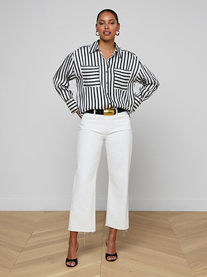 A woman stands with hands on hips, wearing the Eliora Shirt—a relaxed fit striped button-down—paired with white high-waisted cropped pants, a black belt with a gold buckle, black heels, and gold earrings in a minimalist room.