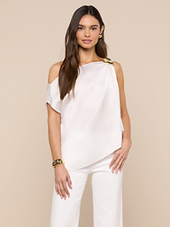 A woman with long brown hair models the Driselle Scarf Top—an off-the-shoulder satin piece with gold hardware—paired with matching white pants, gold earrings, and a bracelet against a beige background.