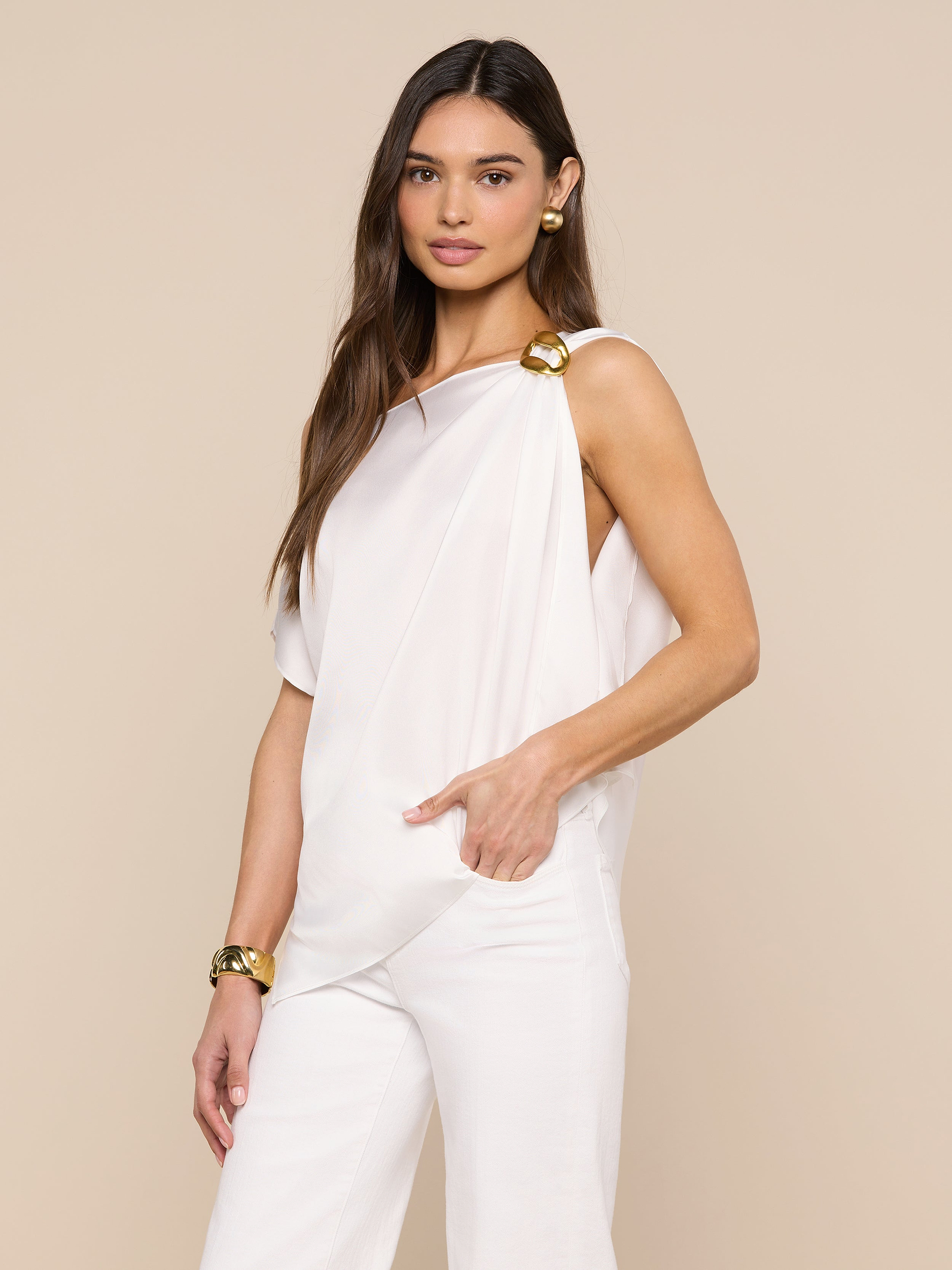 A woman with long brown hair poses against a beige background wearing the Driselle Scarf Top, a sleeveless white satin piece with gold accents on the shoulders, paired with white pants and gold jewelry.