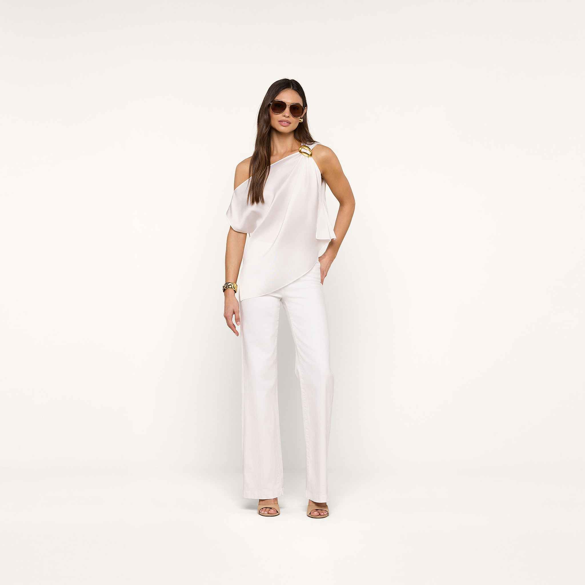 A woman poses confidently against a beige background wearing sunglasses, the Driselle Scarf Top with a gold buckle, white wide-leg pants, gold heels, and a gold bracelet.