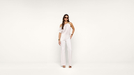 A woman stands against a plain white background wearing a stylish white one-shoulder jumpsuit, sunglasses, bracelets, and sandals.