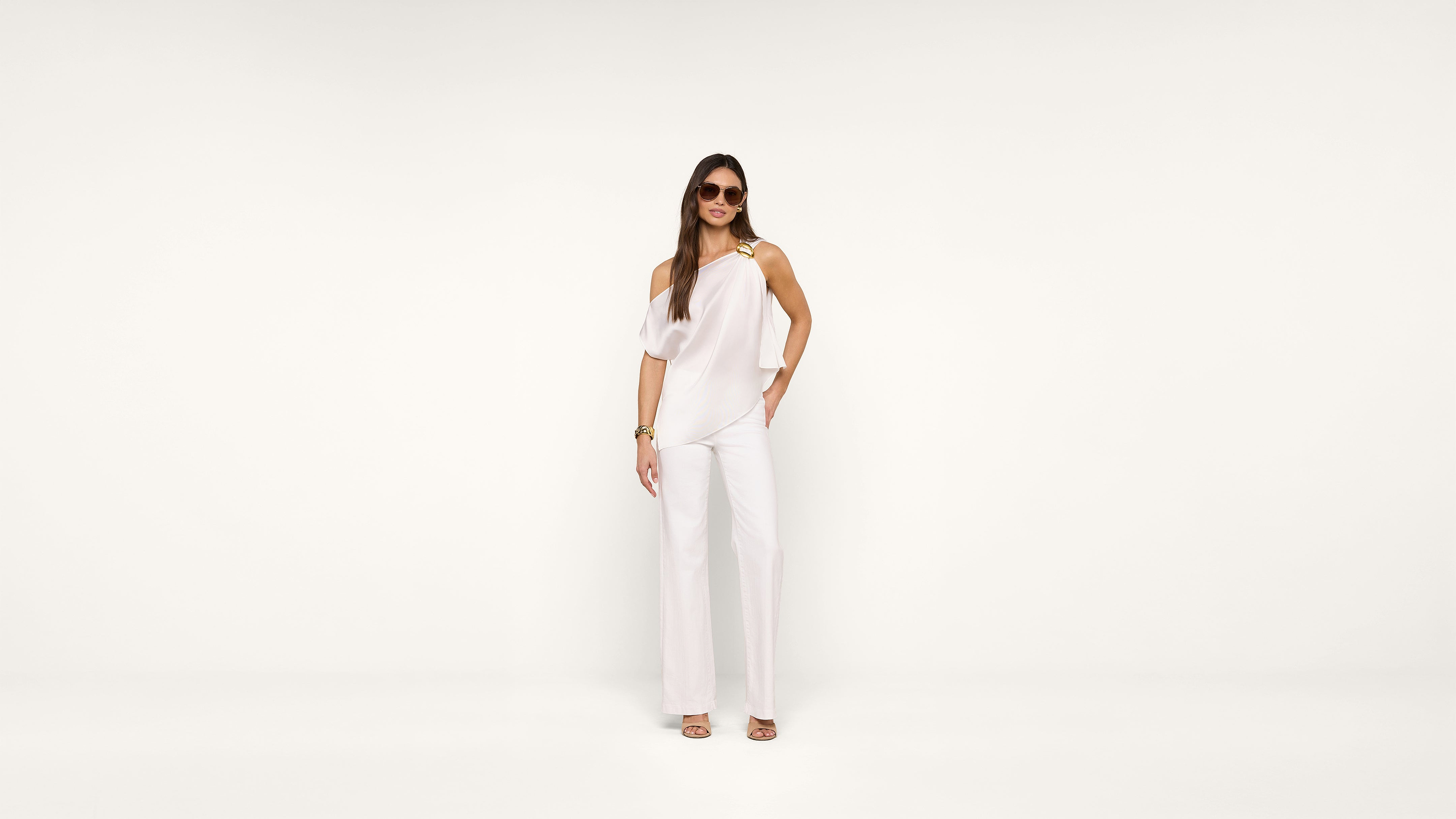 A woman stands against a plain white background wearing a stylish white one-shoulder jumpsuit, sunglasses, bracelets, and sandals.