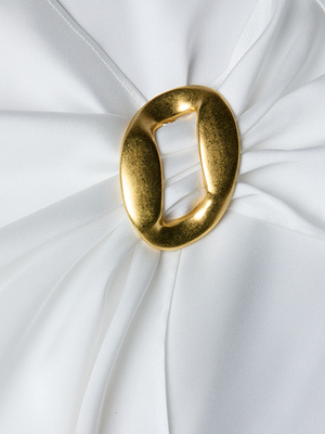 A close-up of the Driselle Scarf Top shows white satin fabric gathered at the center with a shiny, irregularly-shaped gold buckle accent, creating soft, elegant folds that radiate outward.