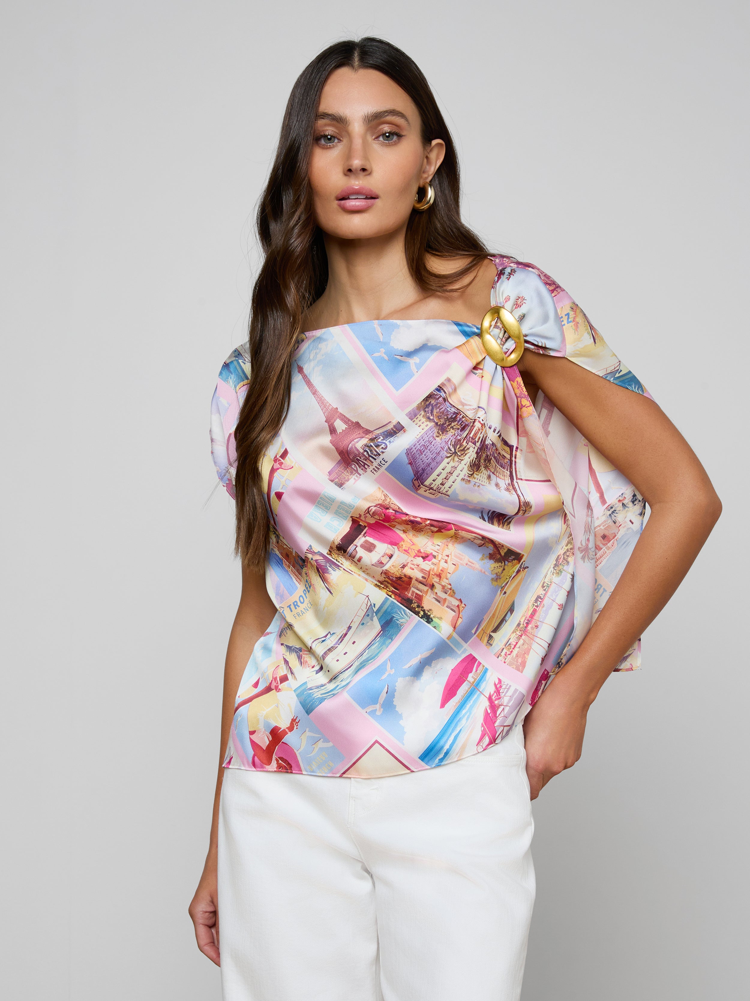 A woman wearing the Driselle Scarf Top, a colorful, patterned short-sleeve blouse, paired with a gold brooch and white pants, poses with one hand in her pocket against a plain background.
