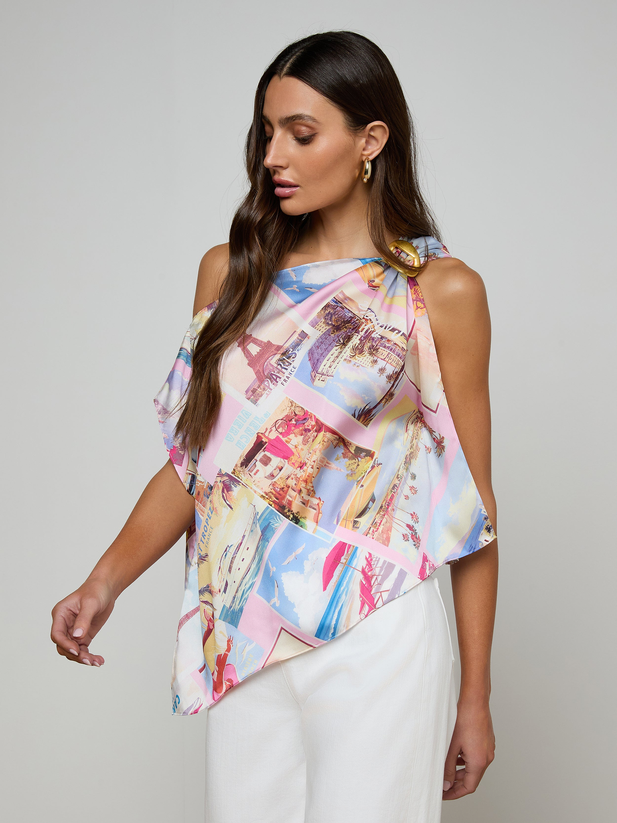A woman wearing the Driselle Scarf Top—a colorful, asymmetrical postcard-print design—paired with white pants, stands against a plain light background, looking down and to the side.