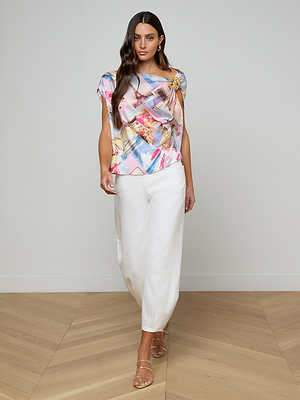 A woman with long brown hair models the Driselle Scarf Top featuring a vibrant postcard print, paired with white wide-leg pants and strappy sandals on a light wooden floor—ideal for vacation style.