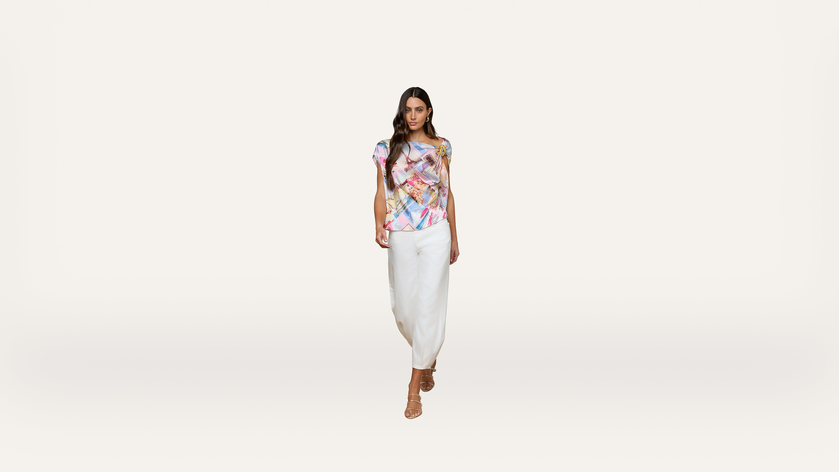 A woman wearing a colorful patterned top, white skirt, and tan heels walks forward against a plain white background.