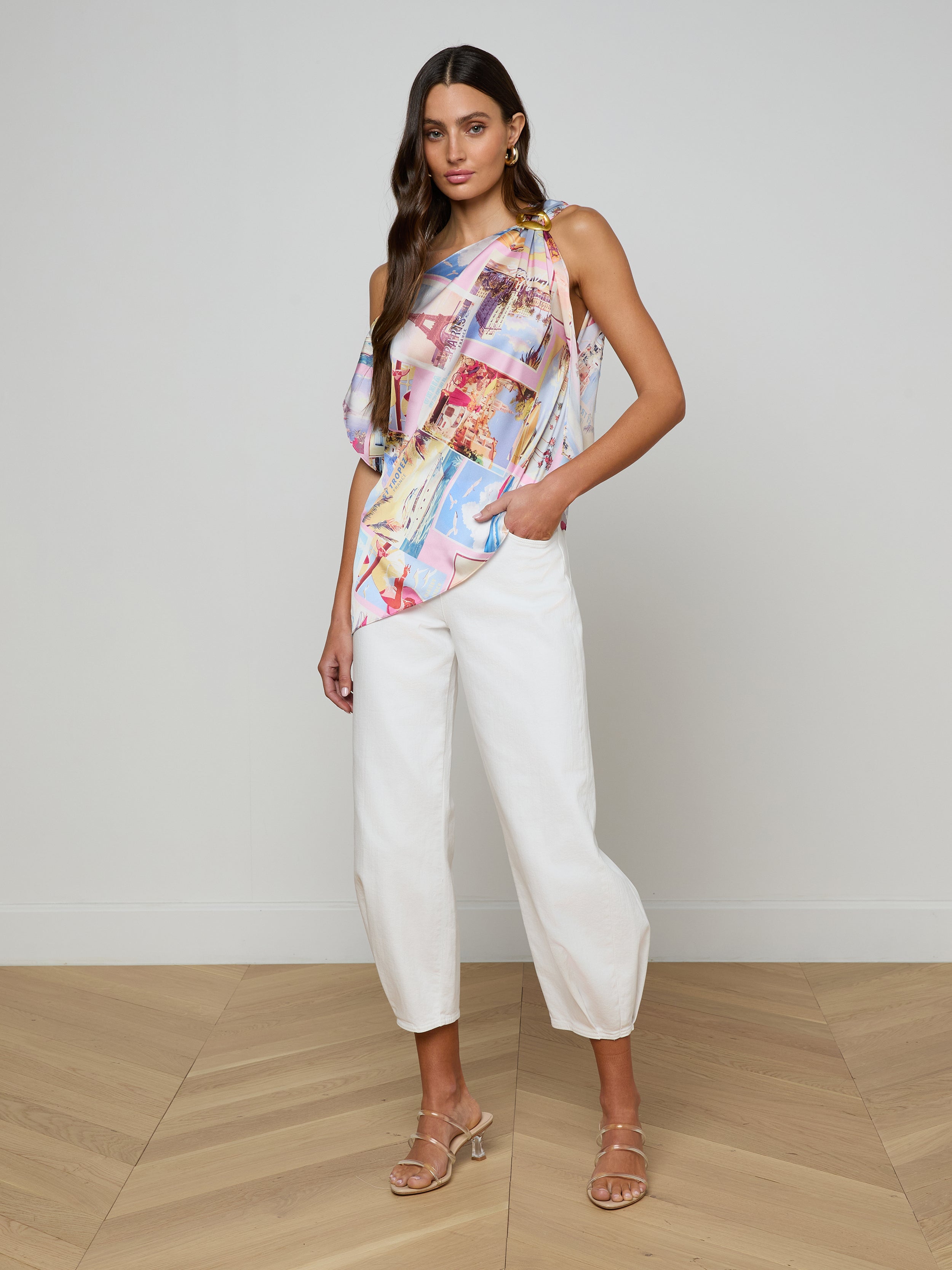 A woman stands against a plain background, wearing the Driselle Scarf Top paired with white cropped pants and beige heeled sandals, one hand in her pocket and the other by her side—an effortless look perfect for vacation wear.
