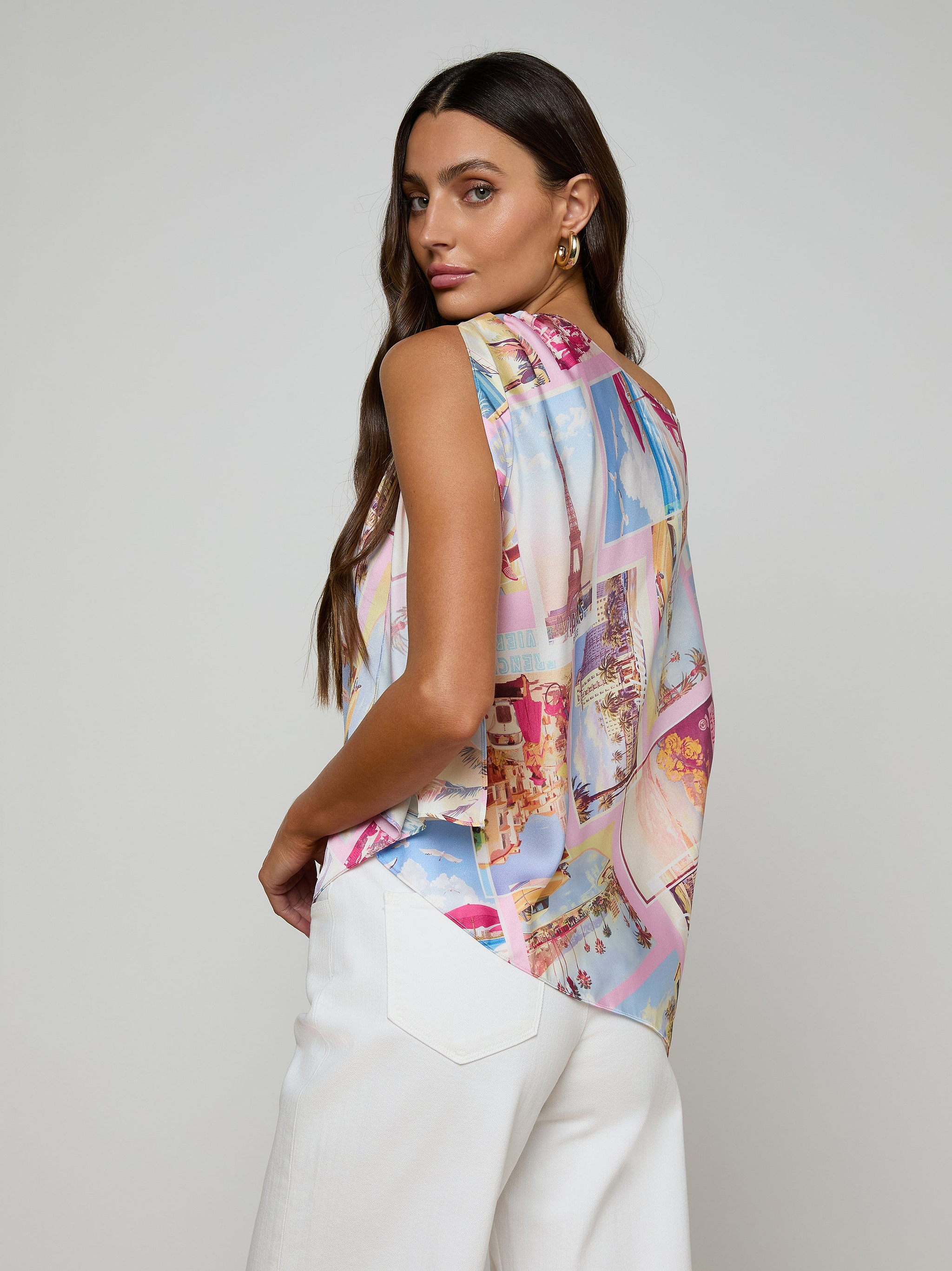 A woman with long brown hair wears the Driselle Scarf Top—a colorful postcard print sleeveless blouse—paired with white pants, looking over her shoulder against a plain light background.