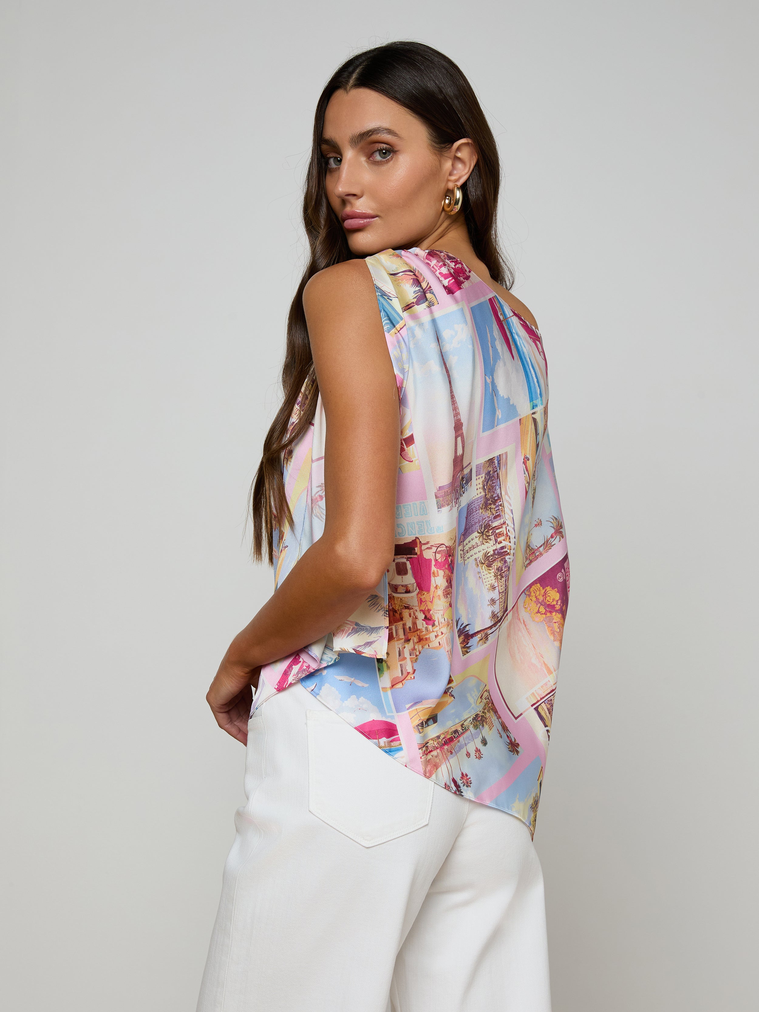 A woman with long brown hair wears the Driselle Scarf Top—a colorful postcard print sleeveless blouse—paired with white pants, looking over her shoulder against a plain light background.