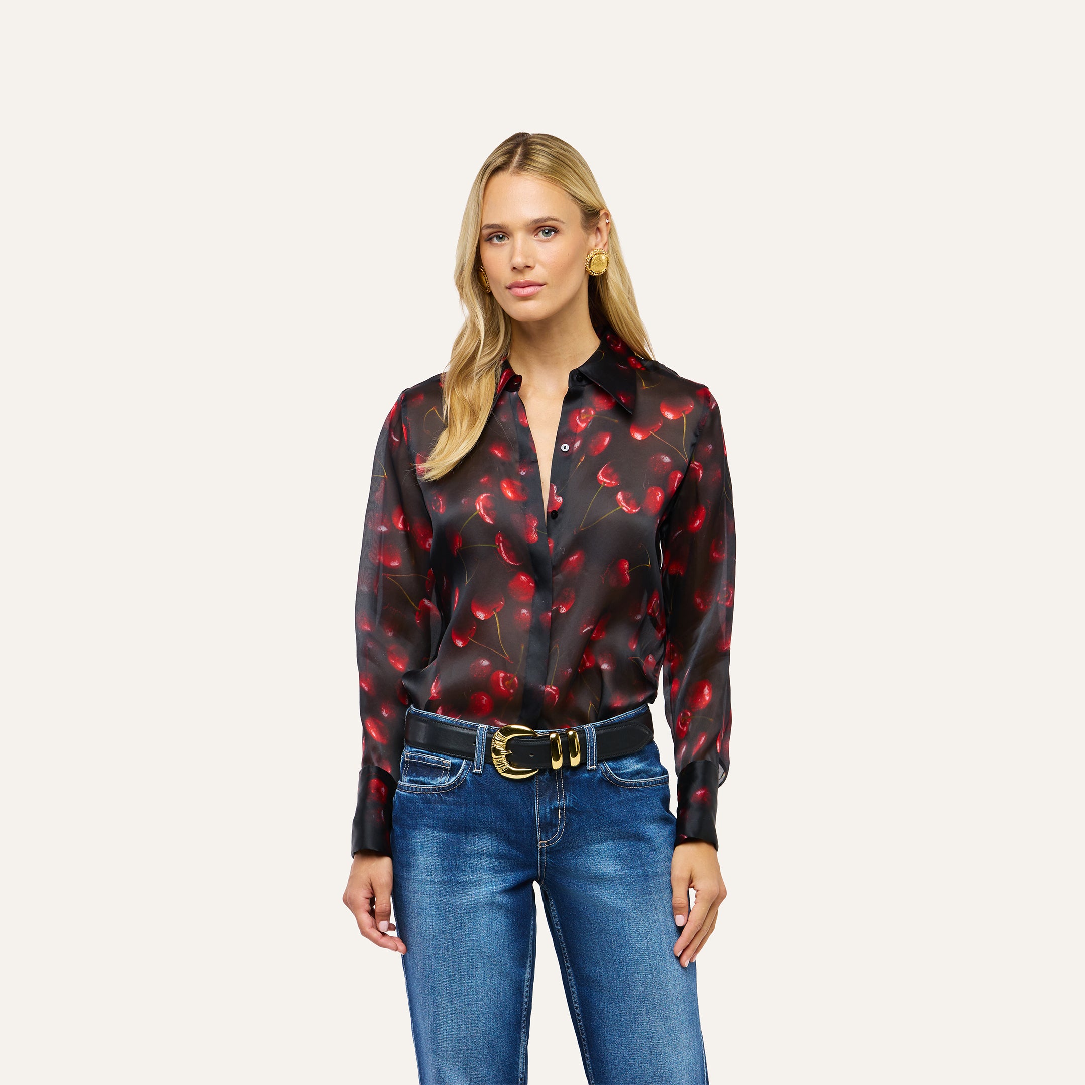 A woman wears a long-sleeved, black button-down Sona Silk Blouse with a cherry print tucked into blue jeans with a black belt.