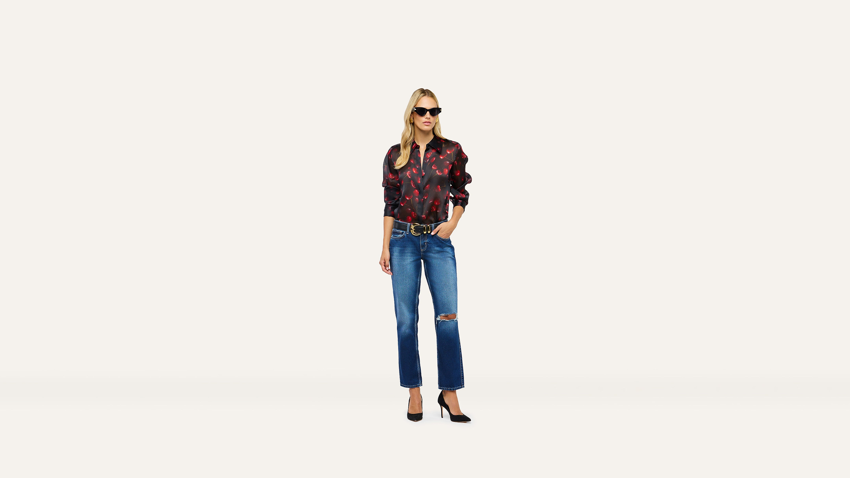 A woman in sunglasses wears a red and black patterned long-sleeve shirt, a gold-buckle belt, and distressed blue jeans with black heels.