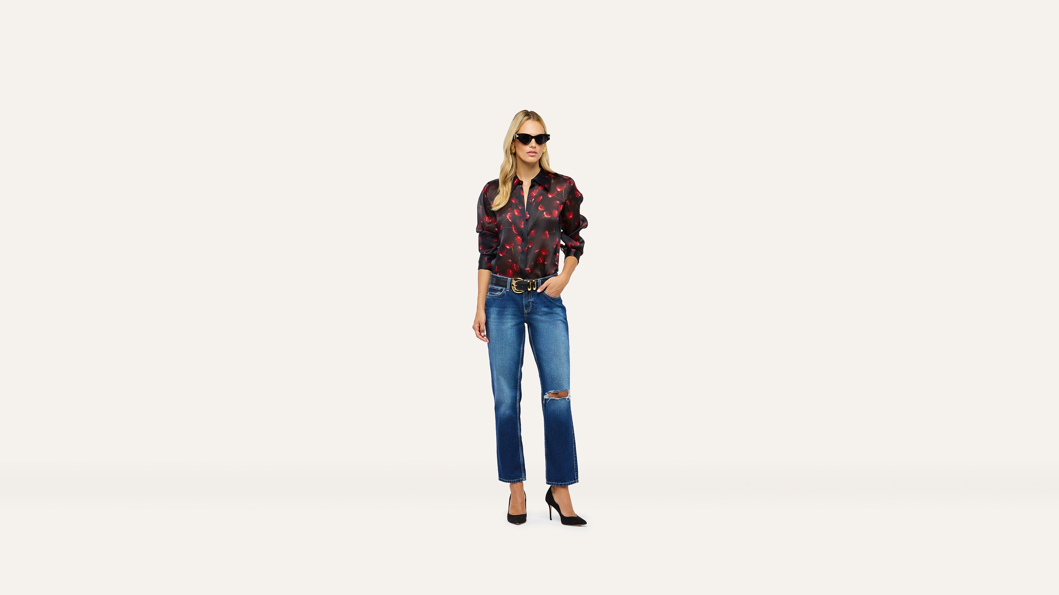 A woman in sunglasses wears a red and black patterned long-sleeve shirt, a gold-buckle belt, and distressed blue jeans with black heels.