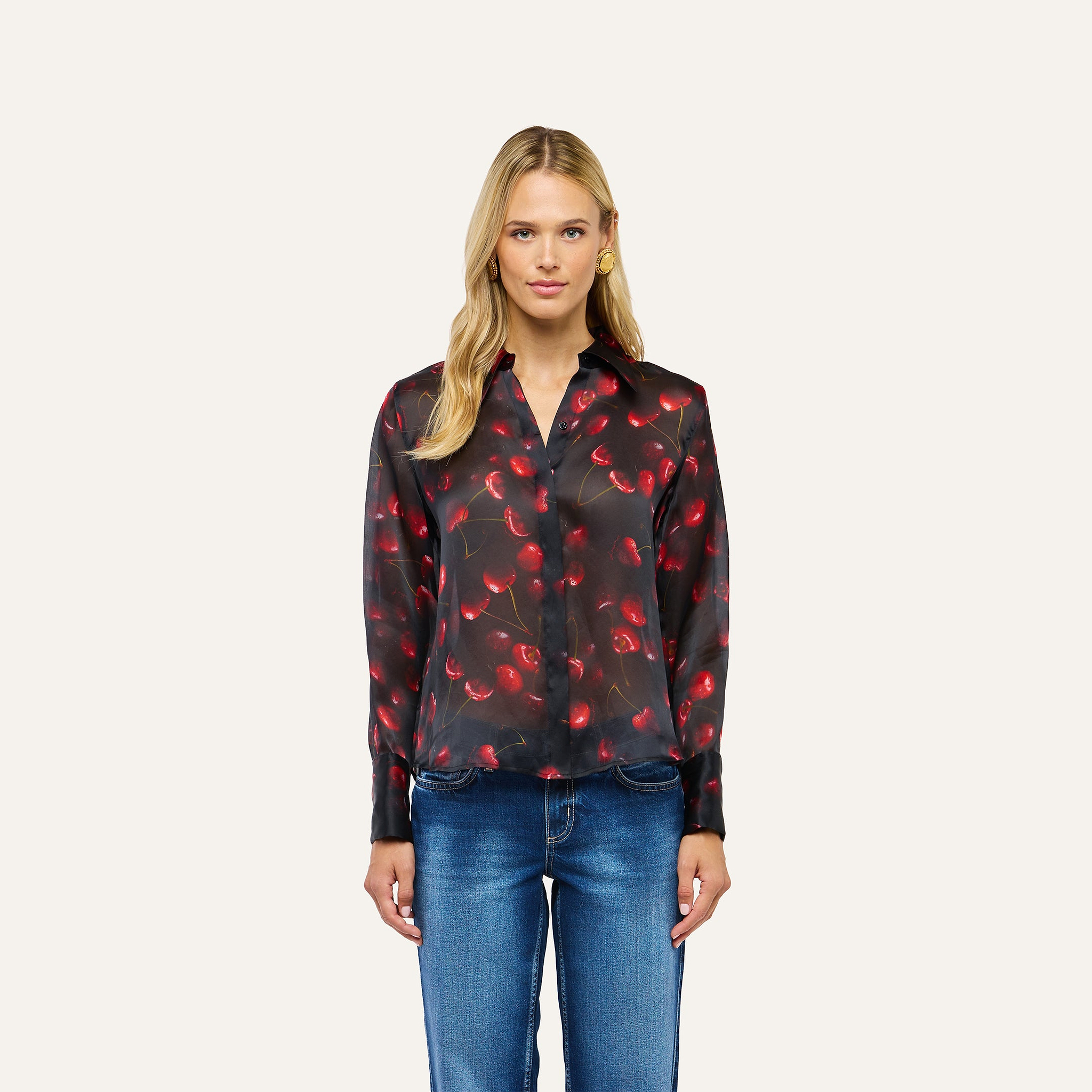 A woman wearing a black Sona Silk Blouse with a red cherry print and blue jeans.