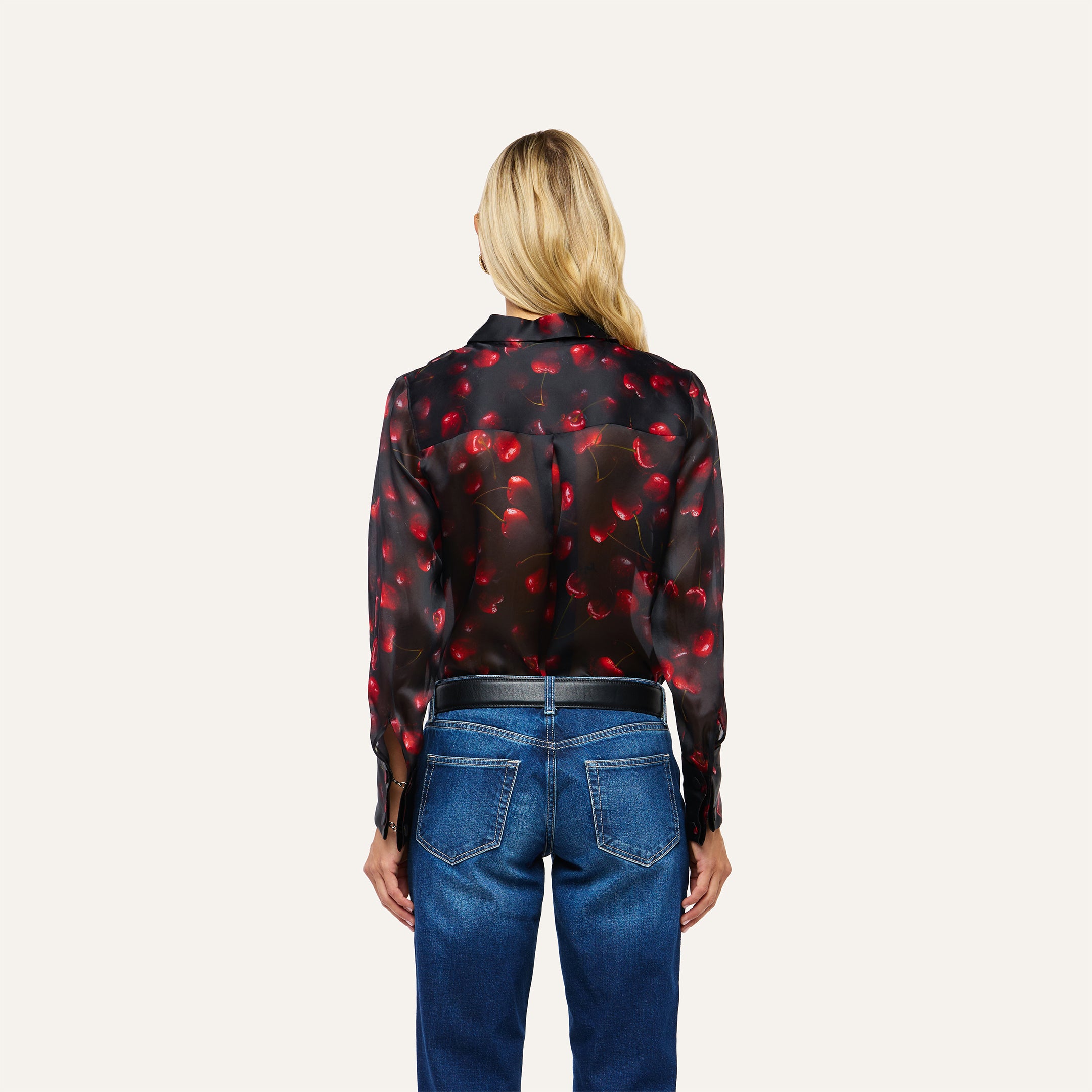 A woman seen from behind wearing a black Sona Silk Blouse with a red cherry print and blue jeans.