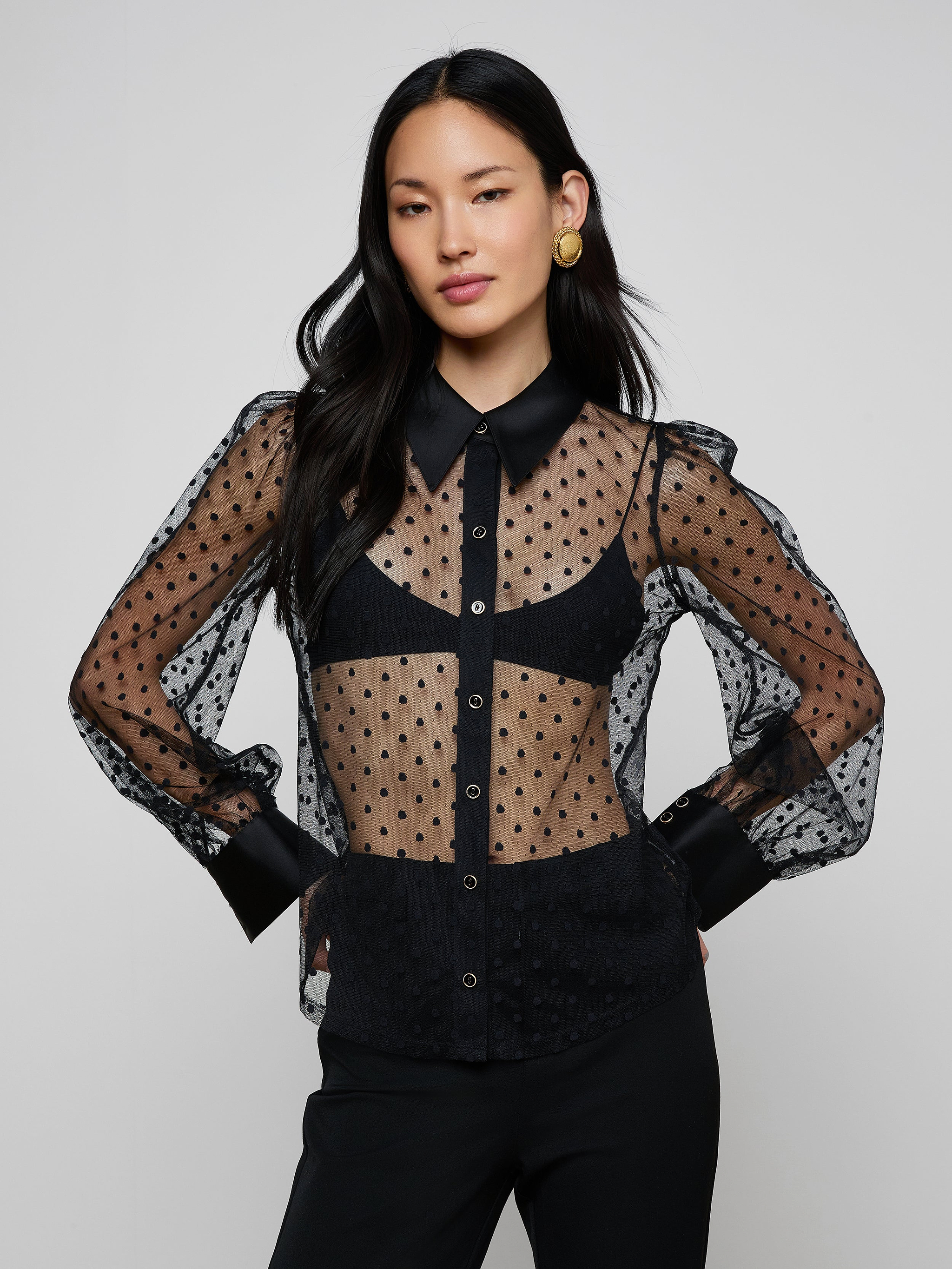 A woman with long dark hair and gold earrings stands against a plain light background, wearing the Sophie Long-Sleeved Blouse—a sheer Swiss dot design—over a black bra, paired with black pants.