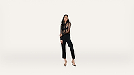 A woman in a sheer black blouse, black pants, and heels stands against a plain white background, looking at the camera with one arm across her waist.