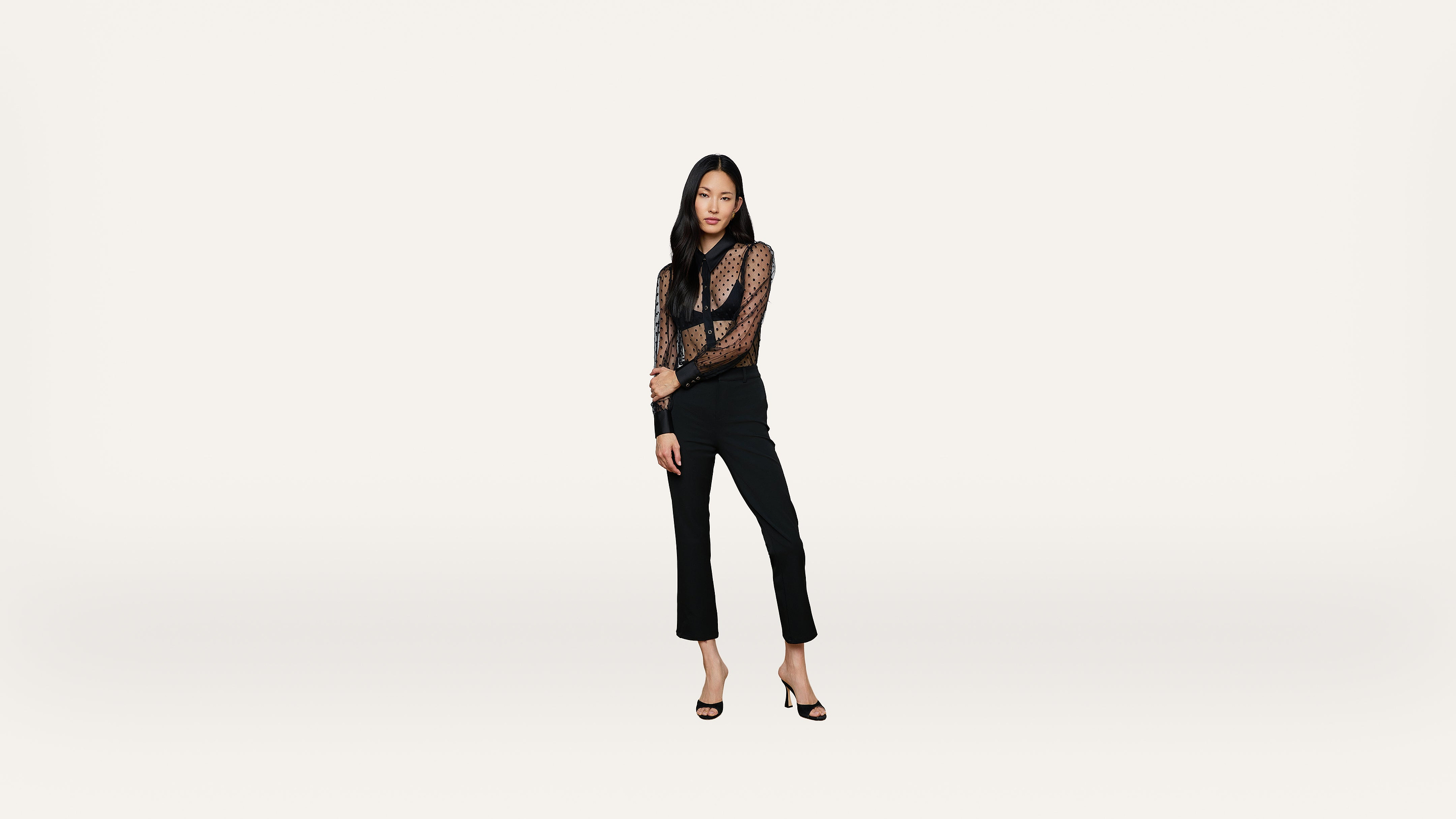 A woman in a sheer black blouse, black pants, and heels stands against a plain white background, looking at the camera with one arm across her waist.