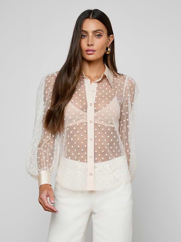A woman with long brown hair wears the Sophie Long-Sleeved Blouse—a sheer, swiss dot, feminine top with puffy sleeves—over a bra and pairs it with white pants, standing against a plain light gray background and looking at the camera.