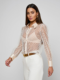 A woman wearing the Sophie Long-Sleeved Blouse—a sheer, swiss dot, feminine top—styled with a nude undergarment, white pants, and a brown belt, stands against a plain light background.