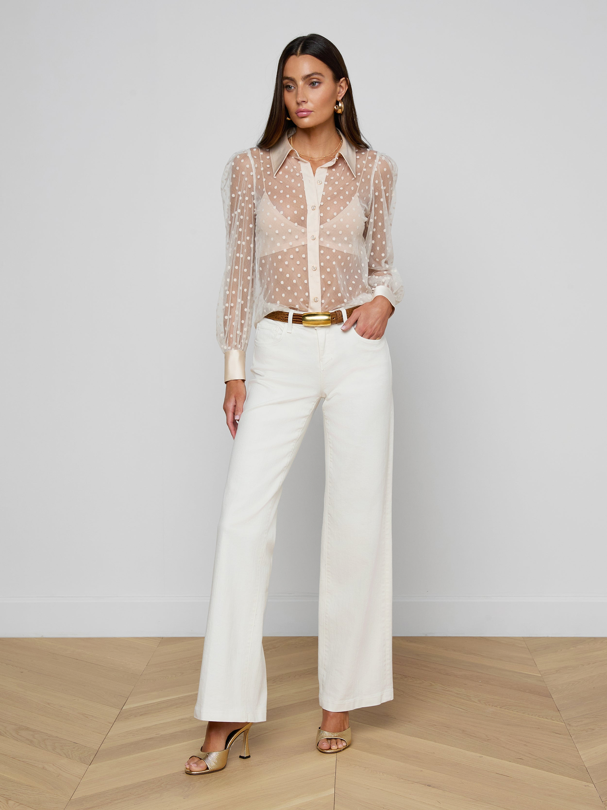 A woman with long hair and a neutral expression stands against a white background, wearing the Sophie Long-Sleeved Blouse—a sheer Swiss dot top—paired with white wide-leg pants, a gold belt, gold earrings, and open-toe heels.