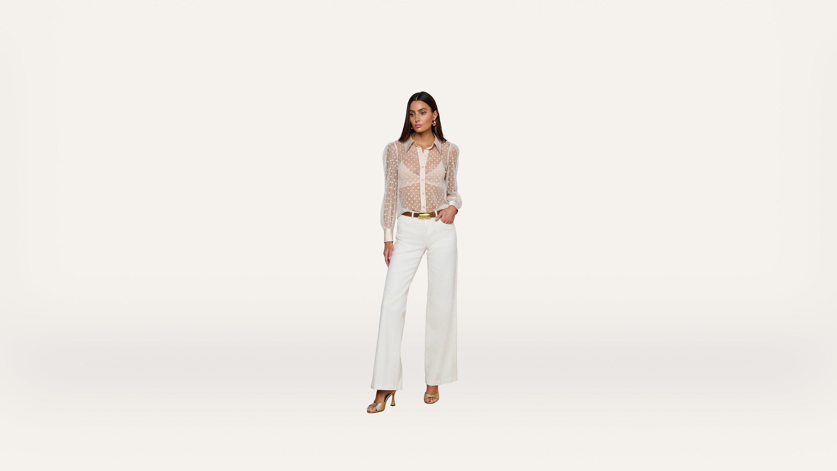 A woman stands confidently in a sheer, polka dot blouse and white wide-leg pants with heels, against a plain, light background.