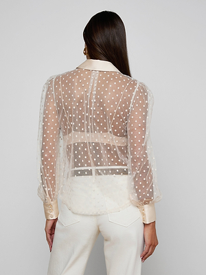 A woman with long brown hair wears the Sophie Long-Sleeved Blouse, featuring sheer swiss dots with satin cuffs and collar, paired with high-waisted white pants. She is turned away, highlighting the blouse’s delicate back detail.