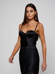 Wearing the Orlina Silk Camisole, a woman with long brown hair poses against a plain light background. The camisole features a fitted black silhouette with thin straps, underwire cups, and delicate lace details.