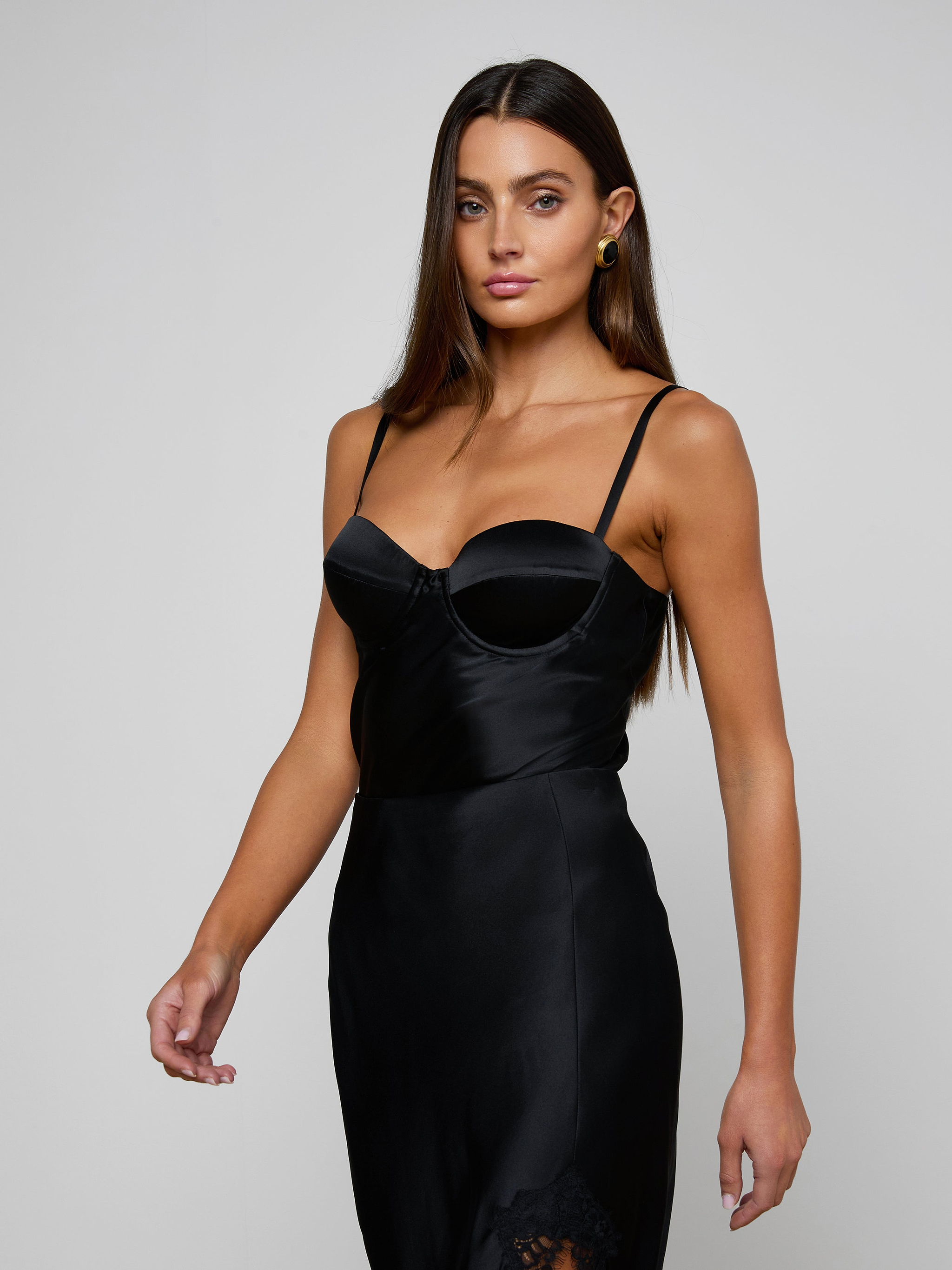Wearing the Orlina Silk Camisole, a woman with long brown hair poses against a plain light background. The camisole features a fitted black silhouette with thin straps, underwire cups, and delicate lace details.