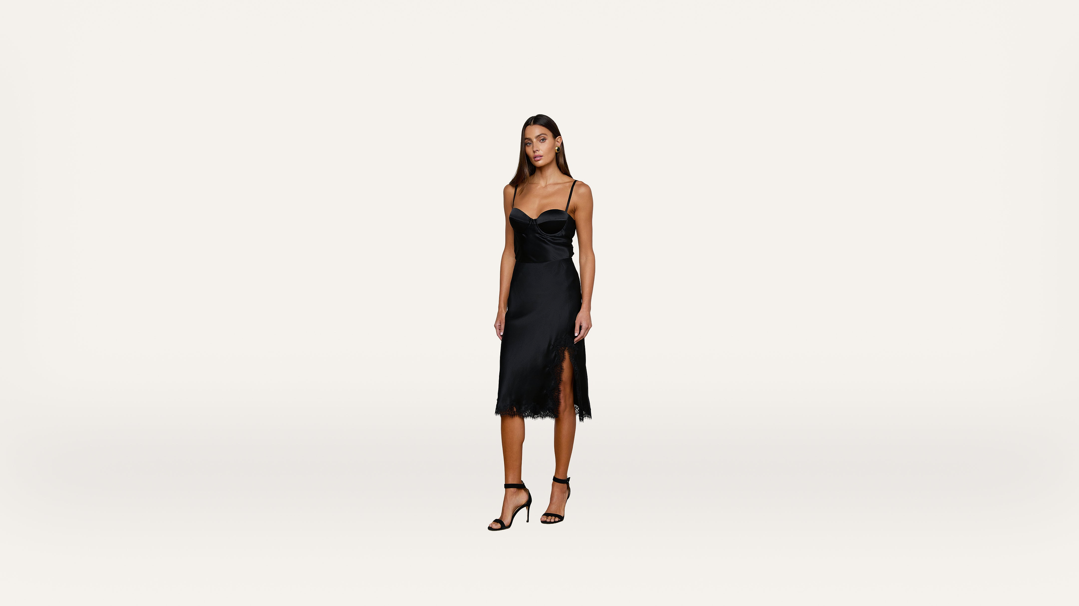 Woman standing and wearing a sleeveless black cocktail dress with a side slit and black high-heeled sandals against a plain white background.