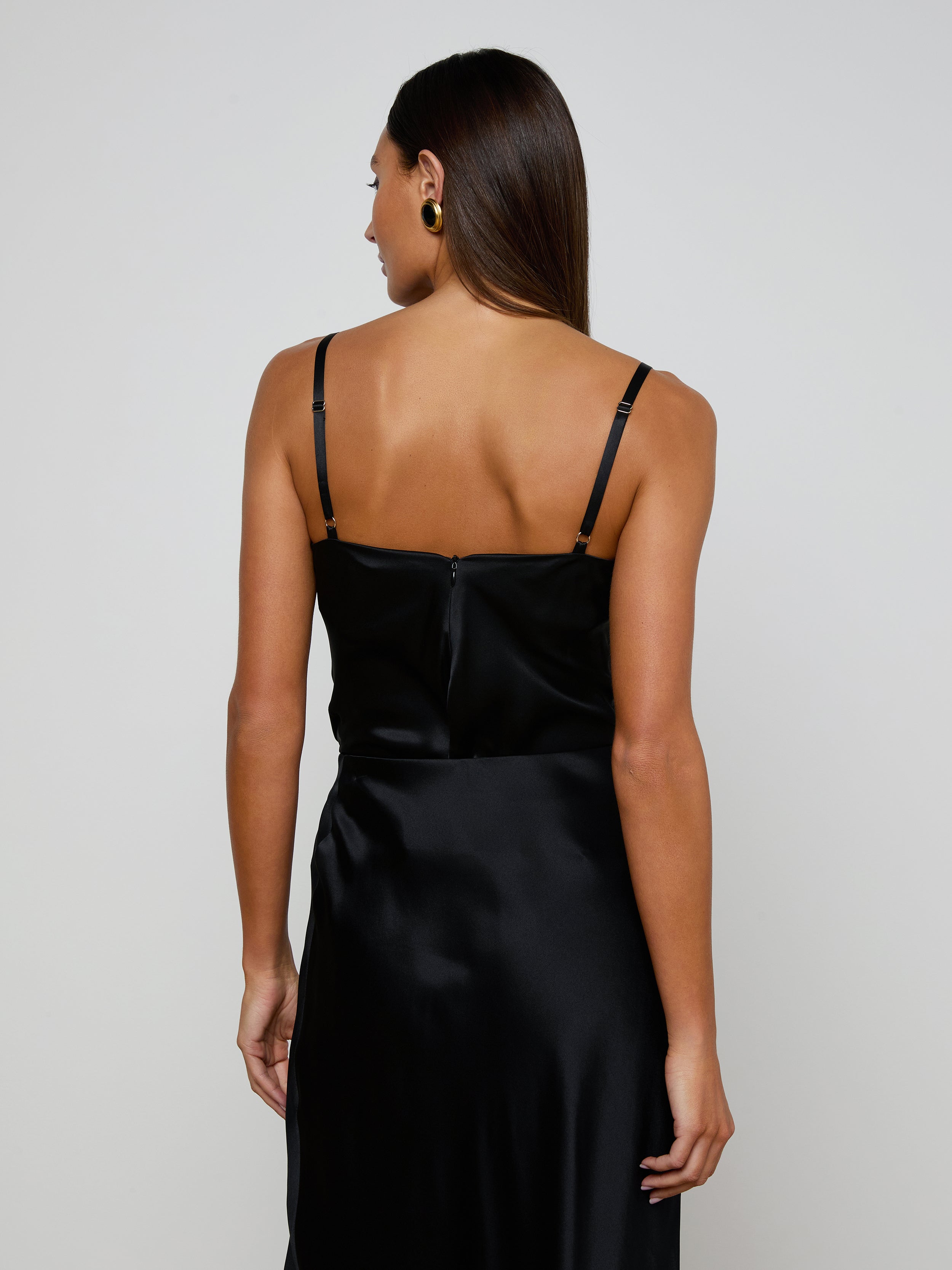 A woman with straight brown hair is seen from behind, standing against a plain light background, wearing the Orlina Silk Camisole in black with thin straps.