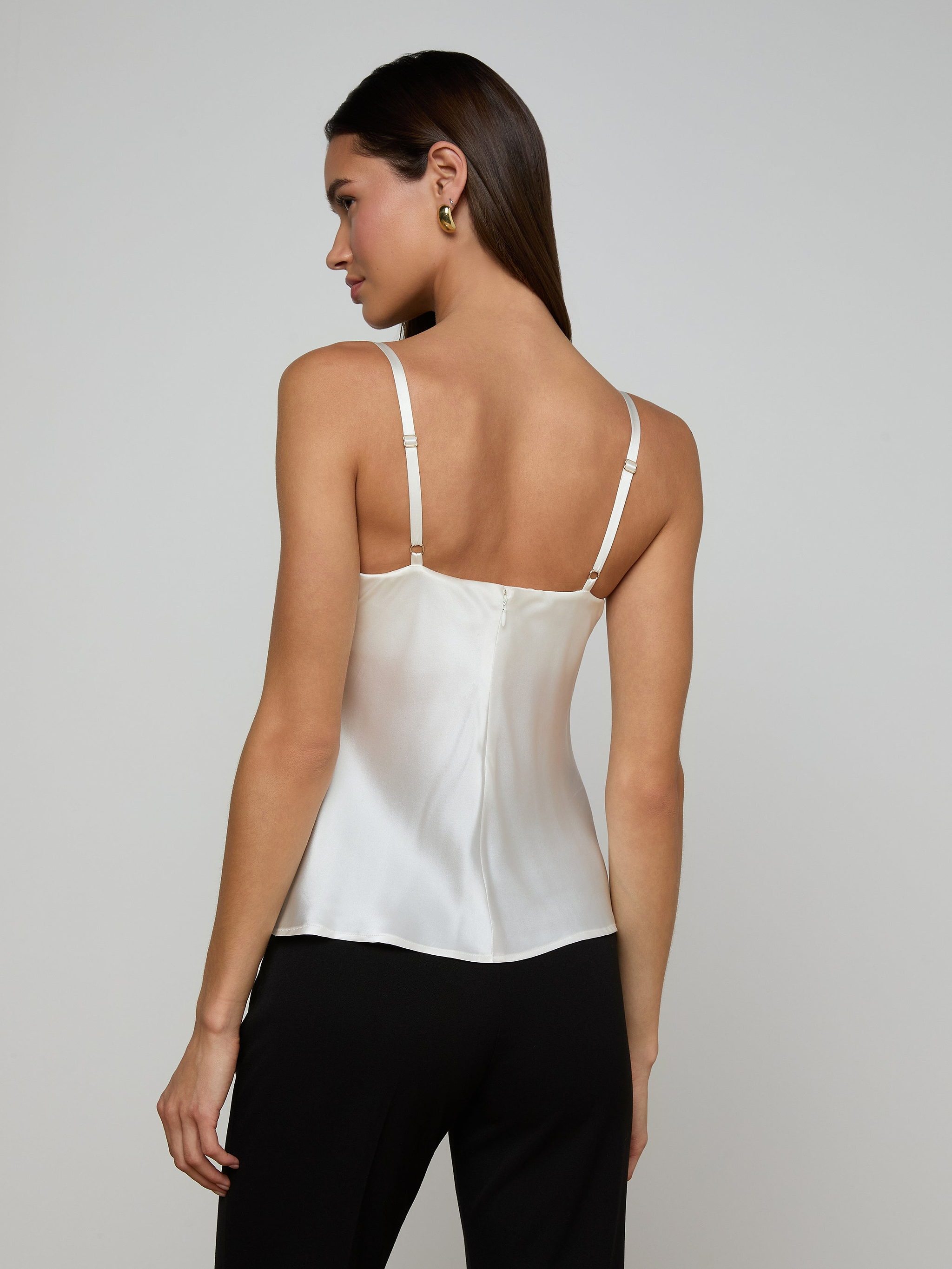 A woman with straight brown hair is shown from the back against a plain light background, wearing the Orlina Silk Camisole in white and black pants. She faces slightly to the left, highlighting the camisole’s adjustable straps and elegant detailing—making it an all-season essential.