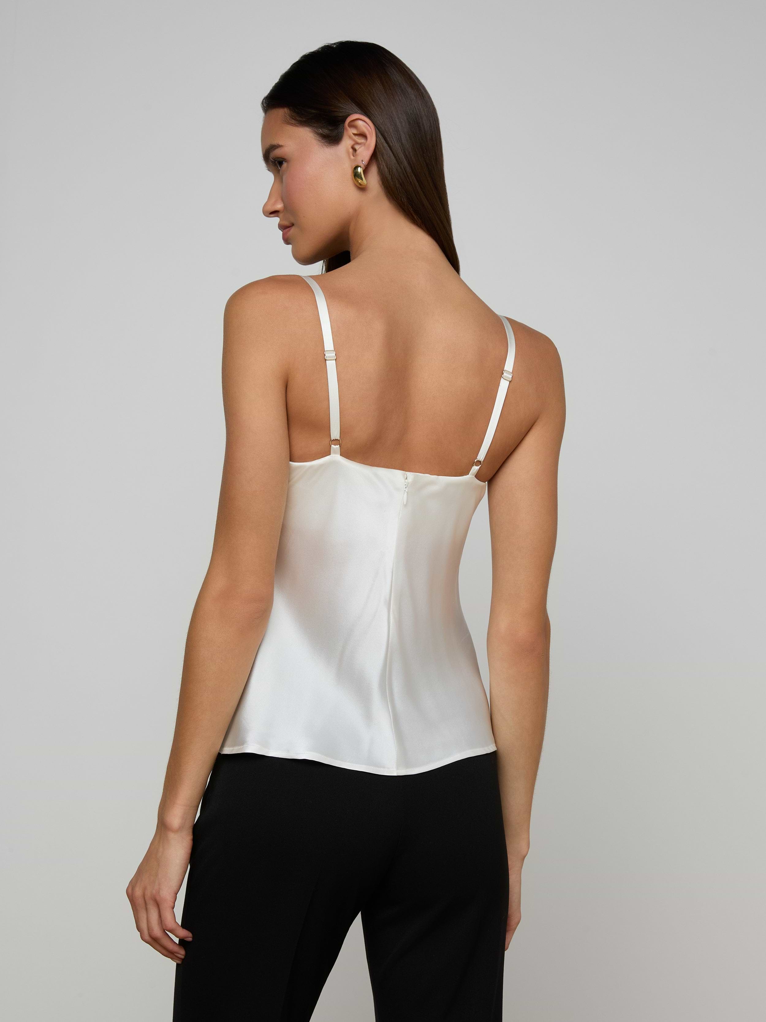 A woman with straight brown hair is shown from the back against a plain light background, wearing the Orlina Silk Camisole in white and black pants. She faces slightly to the left, highlighting the camisole’s adjustable straps and elegant detailing—making it an all-season essential.